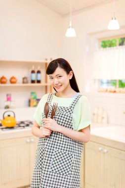 Asyalı kadın mutfak aletlerini elinde tutuyor ve kameraya gülümsüyor.