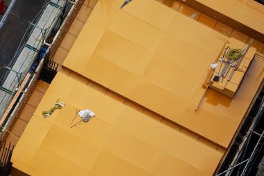 İki inşaat işçisi büyük sarı bina dış yüzeyini boyuyor.