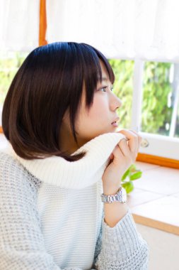 Beyaz kazaklı genç Asyalı kadın pencereden dışarı bakıyor. 