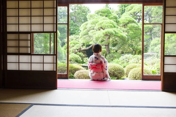 back view of Japanese woman wearing kimono sitting on engawa and looking at garden