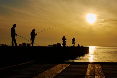 Güneş doğarken deniz kıyısında çubuklarla balık tutan siluetli insanlar.                  