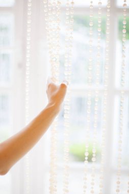Uzanan bir kol ve el, parlak bir pencerede asılı boncuklu bir perdeye nazikçe dokunur..