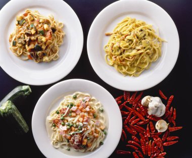 Üç renkli makarna tabağı, beyaz tabaklar ve karanlık bir yüzey üzerinde baharatlar ve sebzeler.