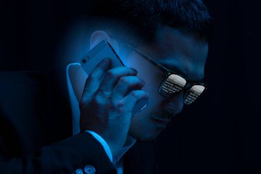 Gözlüklü bilgisayar korsanı karanlık odada akıllı telefon kullanıyor.                          