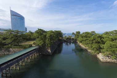 Sakin nehrin yanındaki yemyeşil ağaçların arkasında modern bina yükseliyor.