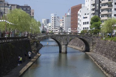 Mavi gökyüzü altındaki şehir binaları arasında sakin bir nehri kaplayan tarihi taş köprü