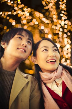 Genç aşık çift gülümsüyor ve Noel ışıklarıyla caddeye bakıyor.    