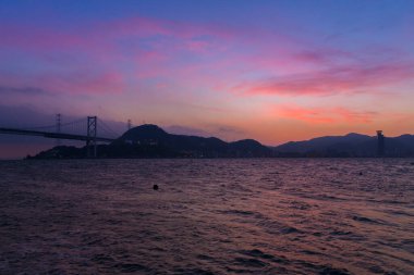 Deniz kıyısında gün batımı, şehir silüeti ve akşamları tepeler. 