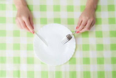 Masanın üzerinde çatal ve bıçak tutan kadın ellerinin üst görüntüsü kareli masa örtüsüyle boş tabağın üzerinde.