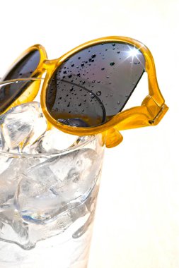 Beyaz zemin üzerinde buz küpleri ve sarı güneş gözlüğü olan bir bardak su, güneşli bir günde yaz tatili ve rahatlama kavramı, kopyalama alanı