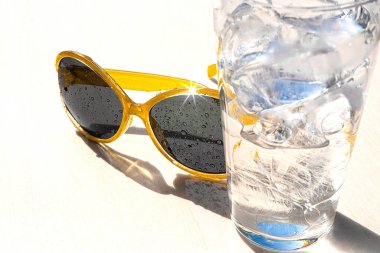Beyaz zemin üzerinde buz küpleri ve sarı güneş gözlüğü olan bir bardak su, güneşli bir günde yaz tatili ve rahatlama kavramı, kopyalama alanı