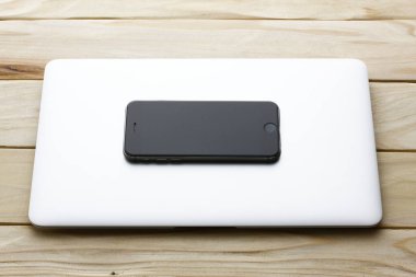 Bir çantada akıllı telefon. Ahşap bir masada dizüstü bilgisayar. Bilgisayarın yanında bir telefon var..