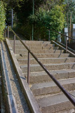 Yeşil çalılar ve metal parmaklıklar arasından yükselen güneşli beton merdivenler.