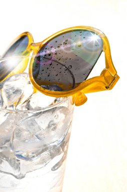 Beyaz zemin üzerinde buz küpleri ve sarı güneş gözlüğü olan bir bardak su, güneşli bir günde yaz tatili ve rahatlama kavramı, kopyalama alanı