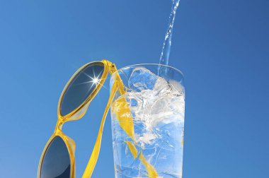 Bir bardak buzlu su ve mavi gökyüzünün arka planında sarı güneş gözlüğü. Güneşli bir günde yaz tatili ve rahatlama kavramı.