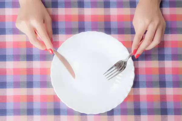 チェッカーテーブルクロス付きテーブル上の白い空のプレートの上にフォークとナイフを保持する女性の手のトップビュー — ストック写真