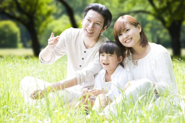 Parkta çimlerin üzerinde oturan küçük kızıyla mutlu Asyalı ebeveynlerin portresi.