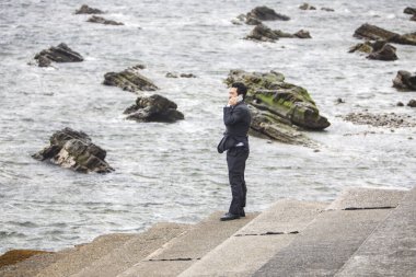 Siyah takım elbiseli Asyalı işadamı deniz kıyısında yürüyor ve telefonla konuşuyor. 