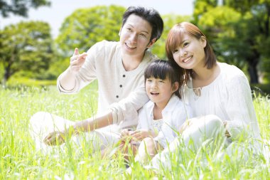 Parkta çimlerin üzerinde oturan küçük kızıyla mutlu Asyalı ebeveynlerin portresi.