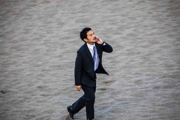 asian businessman in formal suit talking on phone while walking along the sandy beach