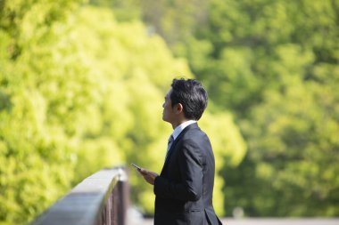 Akıllı telefon kullanan ve parktaki yeşil ağaçlara bakan yakışıklı iş adamının portresi.      