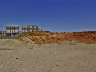 Kuru, kırmızımsı kahverengi toprak ve seyrek bitki örtüsüyle ıssız bir arazi, açık mavi gökyüzünün altındaki apartmanlar.