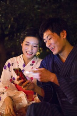 Geleneksel kimono giyen genç Japon çift akşam parkında akıllı telefondan selfie çekiyor.