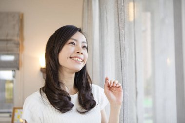 Mutlu genç Asyalı kadın bir evde pencereden dışarı bakıyor.