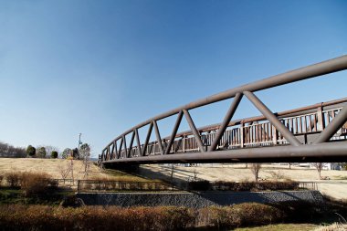 Açık mavi gökyüzünün altında çimenlik alana yayılan metal köprü