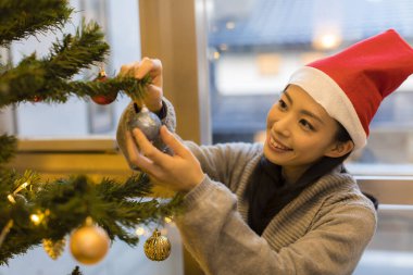 Noel ağacı süsleyen Noel Baba şapkası takan mutlu Asyalı genç kadının portresi