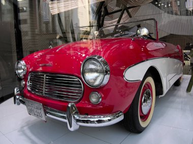 Açık kırmızı ve beyaz renkli üstü açık bir araba müze ortamında sergileniyor..