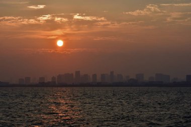 Bir şehrin karanlık silueti gün batımında gökyüzüne yükselir ya da büyük bir su kütlesinin üzerinde güneş doğar..
