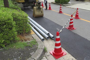 Turuncu trafik konilerinin yanındaki yolun kenarında metal borular var.