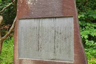 Yeşil yapraklar ve ağaçlarla çevrili eskimiş taş bir anıt.