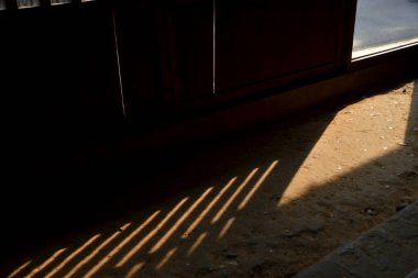 Güneş ışığı şeritleri, loş bir iç mekanda, ahşap bir zeminde doğrusal gölgeler yaratır..