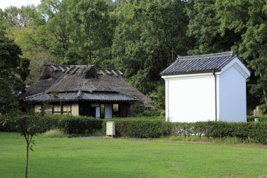 Beyaz binanın yanındaki geleneksel sazdan ev yeşil ağaçların arasında.