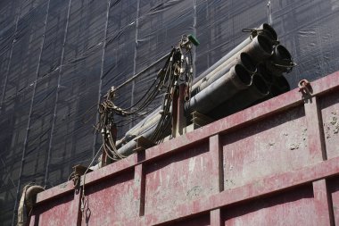 Paslı metal borular dışarıda yıpranmış kırmızı çöp konteynırı duvarında dinleniyor.