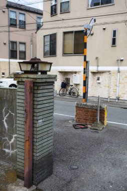 Japonya 'da sokak sahnesi. Fener tarzı aydınlatma armatürüne sahip taş sütun, binanın girişine yakın park edilmiş bir bisiklet, ve kullanım ya da uyarı işareti olabilecek çizgili bir direk.. 