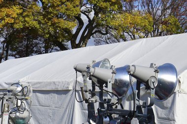 Beyaz olay çadırının yanında duran büyük spot ışıkları sonbahar ağaçları.