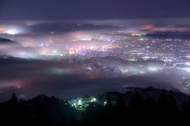 Aydınlanmış şehrin güzel gece manzarası ve sisli karanlık tepeler 