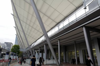 İnsanlar kaldırımda yürüyor. Büyük bir binanın kumaşla kaplı çatısı altında..