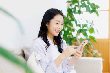 Genç Asyalı kadın oturma odasında akıllı telefon kullanıyor.