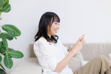Gülümseyen Asyalı kadın evde oturuyor ve cep telefonu kullanıyor.