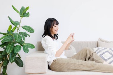 Gülümseyen Asyalı kadın evde oturuyor ve cep telefonu kullanıyor.