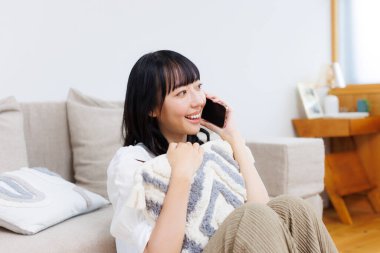 Genç Asyalı güzel kadın evde oturmuş cep telefonuyla konuşuyor. 