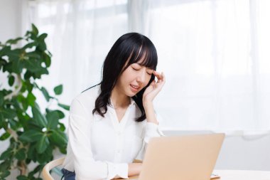 Başı ağrıyan yorgun Asyalı kadın dizüstü bilgisayarla masada oturuyor.