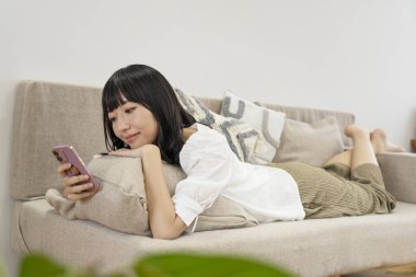 Genç Asyalı kadın oturma odasındaki koltukta akıllı telefon kullanıyor.