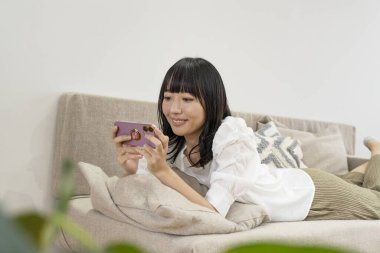 Genç Asyalı kadın oturma odasındaki koltukta akıllı telefon kullanıyor.