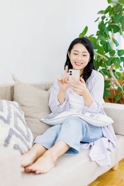 Genç Asyalı kadın oturma odasında akıllı telefon kullanıyor.