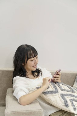 Genç Asyalı kadın oturma odasındaki koltukta akıllı telefon kullanıyor.